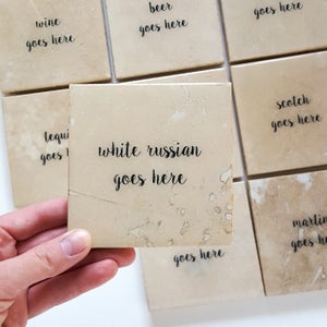 May include: A collection of square coasters with a beige, mottled appearance. One coaster is held up, displaying the text "white russian goes here" in black script. Other coasters read "wine goes here", "beer goes here", and "scotch goes here".