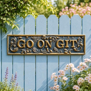 May include: A rectangular wall plaque with a dark textured background and gold-colored raised lettering that reads GO ON GIT. The plaque features an ornate border with sculpted leaf and vine details, and it includes small circular mounting holes.