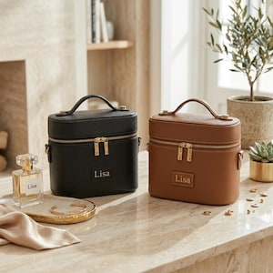 Könnte beinhalten: Zwei personalisierte Kosmetikkoffer, einer schwarz und einer braun, mit goldfarbenen Reißverschlüssen und Griffen. Auf jedem Koffer ist der Name "Lisa" aufgedruckt. Eine Parfümflasche und Schmuck sind ebenfalls zu sehen.