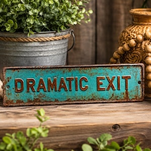 May include: A rectangular metal sign with a distressed turquoise finish and raised, rusted-brown lettering that reads DRAMATIC EXIT. The sign features small circular holes at each end for mounting and exhibits a weathered, aged appearance throughout.