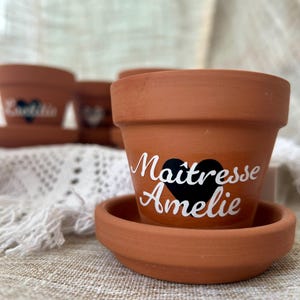 Peut inclure: Petits pots de fleurs en terre cuite avec soucoupes. Un pot porte l'inscription "Maîtresse Amelie" en écriture blanche avec un cœur noir. D'autres pots sont visibles en arrière-plan. Les pots sont posés sur une surface texturée.