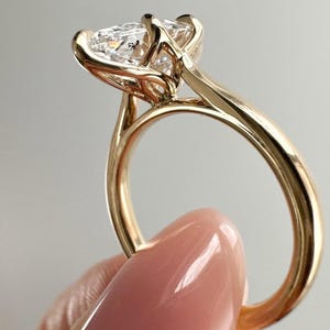 May include: A gold engagement ring with a large, clear, pear-shaped diamond. The ring has a delicate, minimalist design with a thin band and a four-prong setting. The ring is held up against a light gray background.