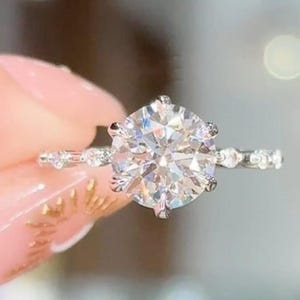 Puede incluir: Un deslumbrante anillo de compromiso con un diamante de talla redonda y gran tamaño. El anillo presenta una banda delicada adornada con piedras de acento más pequeñas, creando un juego de luces. Una joya simbólica de amor y compromiso.