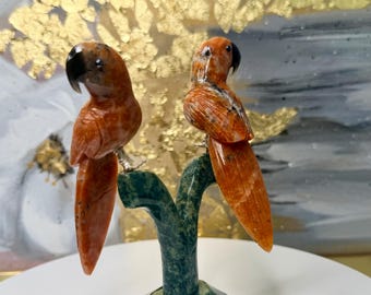 May include: A decorative sculpture featuring two orange parrots perched on a green branch. The parrots have black beaks and are made of a stone-like material. The sculpture is set against a background with gold leaf accents.