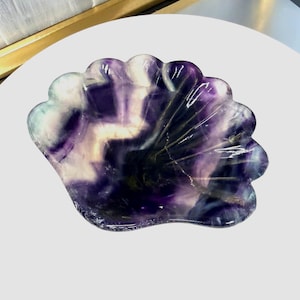 Op de afbeelding: Een decoratieve, schelpvormige schaal gesneden uit een gepolijste, doorschijnende steen. De schaal heeft een wervelend patroon van paarse, witte en heldere tinten. De randen zijn geschulpt en het algehele ontwerp doet denken aan een zeeschelp.