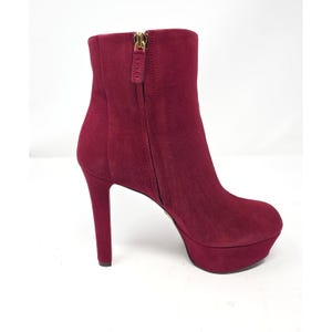May include: A deep burgundy suede ankle boot featuring a high stiletto heel, a thick platform sole, and a gold-toned side zipper with a branded pull tab. The boot has a rounded toe and a smooth, uniform texture across the entire exterior surface.