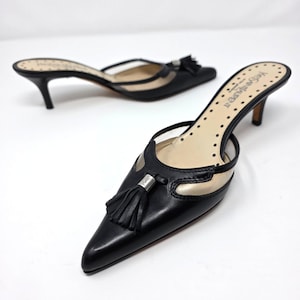 May include: A pair of black leather mules with a pointed toe, featuring a decorative black tassel with a silver-tone metal accent. The shoes have a low kitten heel, clear side panels, and a beige insole with the text Yves Saint Laurent printed.