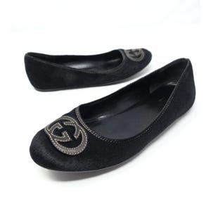May include: A pair of black ballet flats crafted from a textured material, featuring a rounded toe and a prominent interlocking G logo emblem in a metallic grey tone on the front. The shoes have a low profile with visible stitching details.