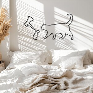 May include: Black metal wall art featuring a cat silhouette and a figure holding an axe. The cat is walking, and the axe figure is standing. The art is mounted on a white wall, with shadows from sunlight. The art is above a bed with white bedding.