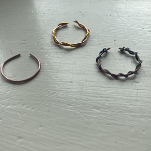 Pode incluir: Três anéis abertos em cobre, ouro e preto. O anel de cobre é uma faixa simples. O anel dourado tem um design torcido. O anel preto tem um design torcido com acabamento escuro. Joias.