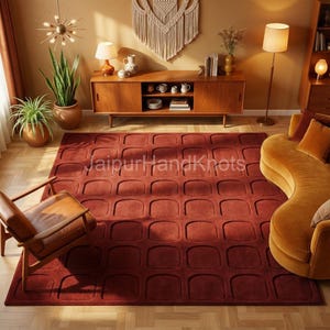 Peut inclure: Un grand tapis rectangulaire de couleur rouille avec un motif géométrique répétitif de carrés arrondis. Le tapis est au centre d'un salon de style moderne du milieu du siècle. Le texte "JaipurHandKnots" est visible sur le tapis.