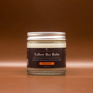 Op de afbeelding: Een glazen pot Holistic Heritage Tallow Bee Balm met een zilveren deksel. Het etiket luidt "Tallow Bee Balm" en "Citrus Grove". De pot bevat een romige, crèmekleurige balsem. De tekst vermeldt ook Iers grasgevoerd biologisch rundvet.