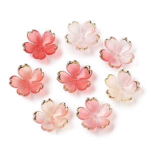 May include: Eight pink and white flower-shaped resin embellishments with gold trim. The embellishments are perfect for adding a touch of elegance to crafts and jewelry.