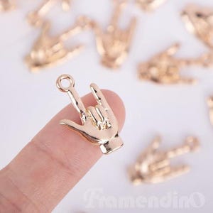 May include: Gold-colored metal charm in the shape of a hand making the "I love you" sign. The charm has a small loop at the top for attaching to jewelry or other crafts. Several similar charms are scattered in the background.