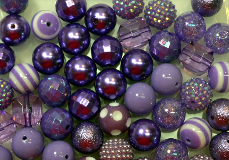 May include: A collection of purple beads in various shapes and textures. The beads include faceted, striped, and polka-dotted designs. Some beads have a shiny, metallic finish, while others are matte. The color palette ranges from light lavender to deep violet.