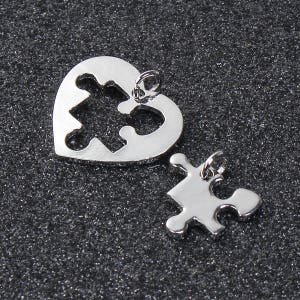 May include: Two silver-colored pendants. One is heart-shaped with a puzzle piece cutout. The other is a puzzle piece. Both have small loops for attaching to a chain or cord. The pendants are on a dark, textured surface.