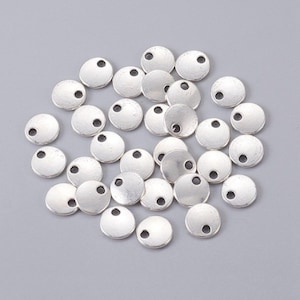 May include: A collection of silver-toned metal circle charms with a small hole at the top. The charms are scattered on a gray surface.