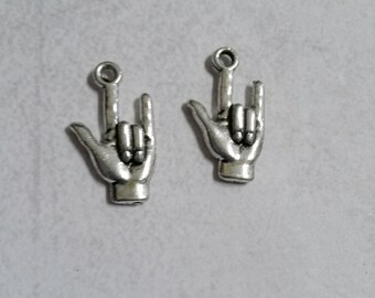 Puede incluir: Dos colgantes de metal plateado con forma de manos haciendo el signo "Te quiero". Cada colgante tiene un lazo en la parte superior para sujetarlo a joyas u otras manualidades. Los colgantes están sobre un fondo gris claro.