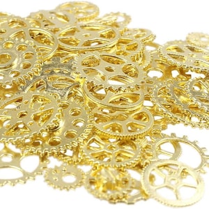 May include: A collection of small, gold-colored metal gears of various sizes, scattered against a white background. The gears have intricate designs with multiple spokes and circular holes, suggesting a steampunk or industrial theme.