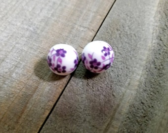 May include: Two white, round beads with a floral pattern in shades of purple. The flowers are clustered around the bead's surface. The beads are displayed on a weathered, gray wooden surface.