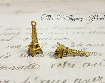 May include: Two gold-tone Eiffel Tower charms. The charms are detailed and have a vintage look. The charms are on a white background with handwritten text that reads "The Slippery Pearl".