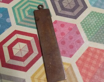 May include: A rectangular, bronze-colored metal pendant with a small loop at the top. The pendant is lying on a colorful patterned paper with umbrella designs in various colors, including red, blue, and yellow.