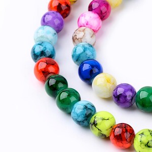 May include: Close-up of a strand of colorful, round beads in various shades including red, green, blue, purple, and yellow. The beads have a marbled or speckled appearance, and are strung together on a white background. The beads are approximately 1 cm in diameter.