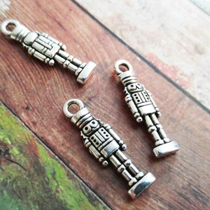May include: Three silver-coloured metal charms shaped like toy soldiers. Each charm has a loop at the top for attaching to a necklace or bracelet. The charms are on a wooden surface with brown, green, and orange tones.