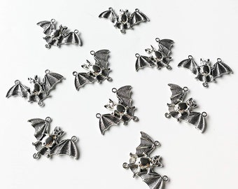 May include: Silver-colored bat charms with detailed wing designs. Each charm features a central body with a small, round embellishment. The charms are scattered across a white surface, ideal for jewelry making or crafting projects.