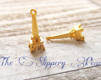 May include: Two gold-coloured Eiffel Tower charms, one upright and one lying down, are displayed on a light-coloured, textured surface. The charms are detailed with architectural features. The text "The Slippery Pearl" is written in cursive.