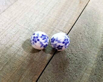 May include: Two white ceramic beads with a blue floral pattern. The beads are round and have a glossy finish. The blue floral design includes small flowers and leaves. The beads are displayed on a wooden surface.