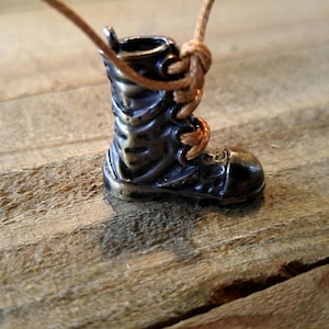 May include: A small, bronze-toned boot pendant with detailed lacing and a loop at the top. It hangs from a tan cord, possibly a necklace or charm. The boot is displayed on a rustic wooden surface, adding to its charm and appeal.