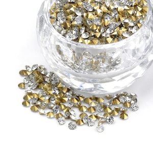 Op de afbeelding: Een glazen kom gevuld met heldere en gouden strass steentjes. De strass steentjes zijn verspreid over een wit oppervlak.