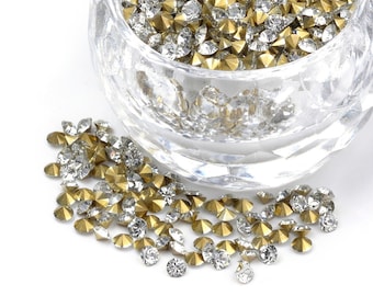 May include: A glass bowl filled with clear and gold rhinestones. The rhinestones are scattered on a white surface.
