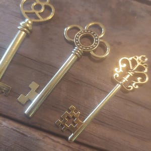 Può includere: Tre chiavi scheletriche dorate ornate con disegni intricati sulle teste delle chiavi. Le chiavi sono disposte su una superficie di legno.