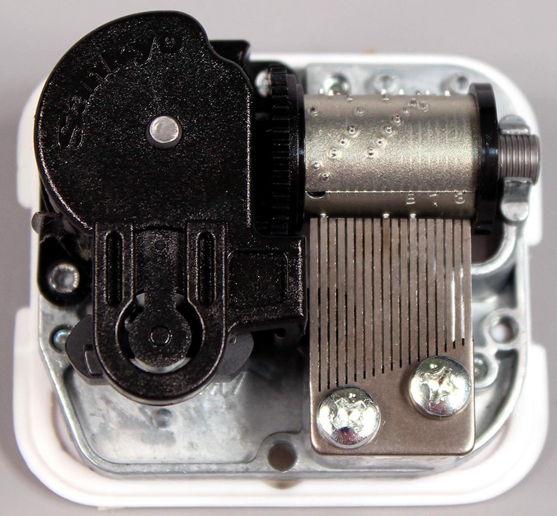 May include: A close-up view of a silver and black music box mechanism. The mechanism has a winding key, a metal comb, and a silver cylinder with holes punched into it. The mechanism is mounted on a white plastic base.