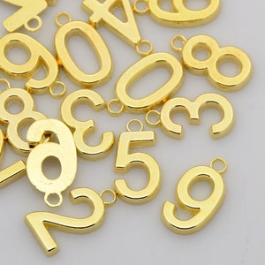 May include: A collection of gold-colored metal number charms. The numbers include 0, 1, 2, 3, 4, 5, 6, 7, 8, and 9. Each charm has a small loop at the top for attaching to a necklace, bracelet, or other jewelry.