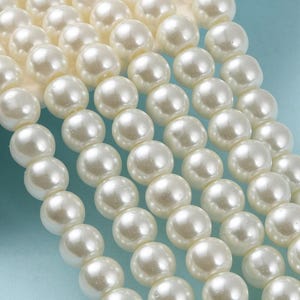 May include: Close-up of multiple strands of creamy white faux pearls. The pearls are round, smooth, and have a subtle luster. The beads are arranged in neat rows, suggesting they are ready for jewelry making or crafting.