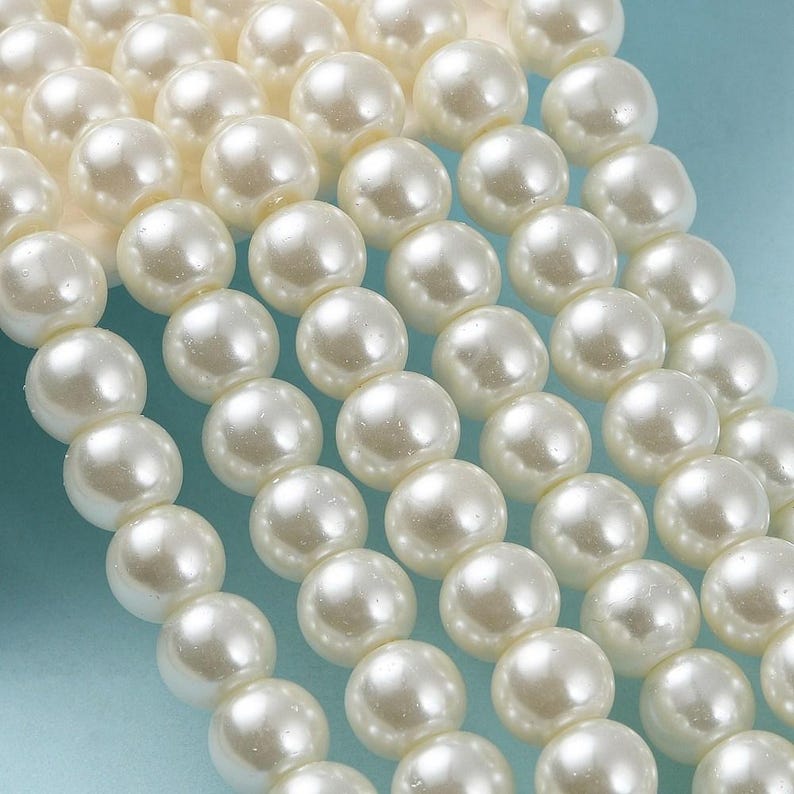 May include: Close-up of multiple strands of creamy white faux pearls. The pearls are round, smooth, and have a subtle luster. The beads are arranged in neat rows, suggesting they are ready for jewelry making or crafting.