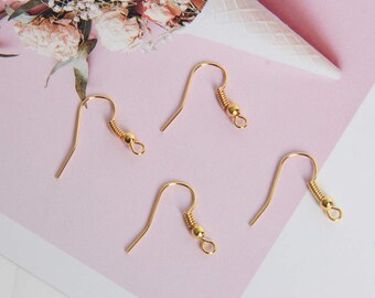 May include: Close-up of five gold-colored earring hooks. Each hook has a curved wire design with a coiled spring and a small ball at the top, and a loop at the bottom. The hooks are arranged on a pink and white surface.
