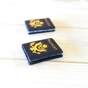 May include: Two miniature blue passports with the word "PASSPORT" printed in gold on the cover. The passports are on a white wooden surface.