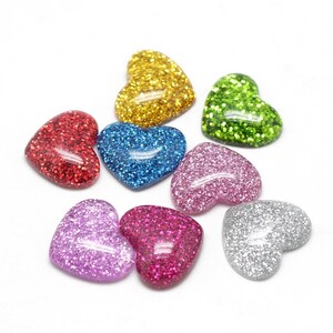 Op de afbeelding: Een verzameling glitterende hartvormige versieringen in verschillende kleuren, waaronder rood, goud, blauw, groen, roze, paars en zilver. Deze decoratieve harten zijn verspreid tegen een witte achtergrond.