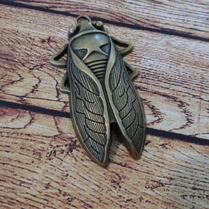 Op de afbeelding: Een bronzen metalen cicade hanger met gedetailleerde vleugel- en lichaamskenmerken. De insectvormige hanger heeft een lus aan de bovenkant om op te hangen en is geplaatst tegen een houten achtergrond.