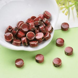 May include: A collection of red and brown ceramic beads with a speckled pattern. The beads are cylindrical and round, with a hole through the center. Some beads are in a white bowl, while others are scattered on a green surface.