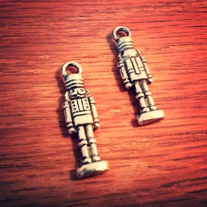 May include: Two silver-toned nutcracker charms with a detailed design. The charms have a loop at the top for hanging.