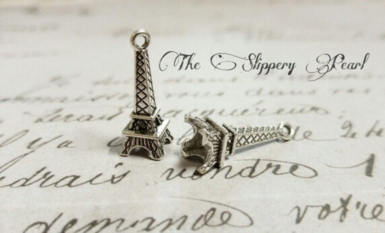 May include: Two silver Eiffel Tower charms, one standing upright and the other lying on its side. The charms are on a white background with cursive writing.