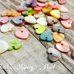 Puede incluir: Una colección de cuentas en forma de corazón en varios colores, incluyendo rosa, amarillo, verde y gris, dispersas sobre una superficie de madera envejecida. Cada cuenta tiene un pequeño agujero para ensartar. El texto "The Slippery Pearl" está abajo.