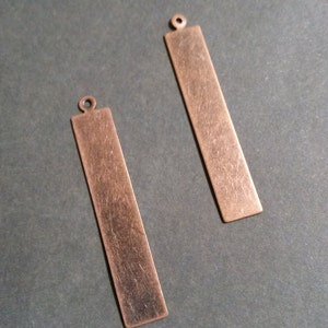 Puede incluir: Dos colgantes rectangulares de metal color cobre con acabado cepillado. Cada colgante tiene un pequeño lazo en la parte superior para sujetarlo a la joyería. Los colgantes se muestran sobre una superficie gris oscuro.