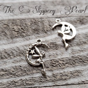May include: Two silver-colored fairy charms. Each charm features a fairy silhouette perched on a crescent moon with stars. The charms have a loop at the top for attaching to jewelry. The text "The Slippery Pearl" is at the top.