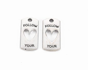 Puede incluir: Dos etiquetas de amuleto plateadas con las palabras "Follow Your" y un corazón recortado en el centro.